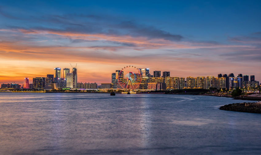 Illuminated buildings by sea against sky during sunset