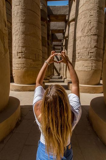 Rear view of woman making hand heart shape while standing amidst architectural column
