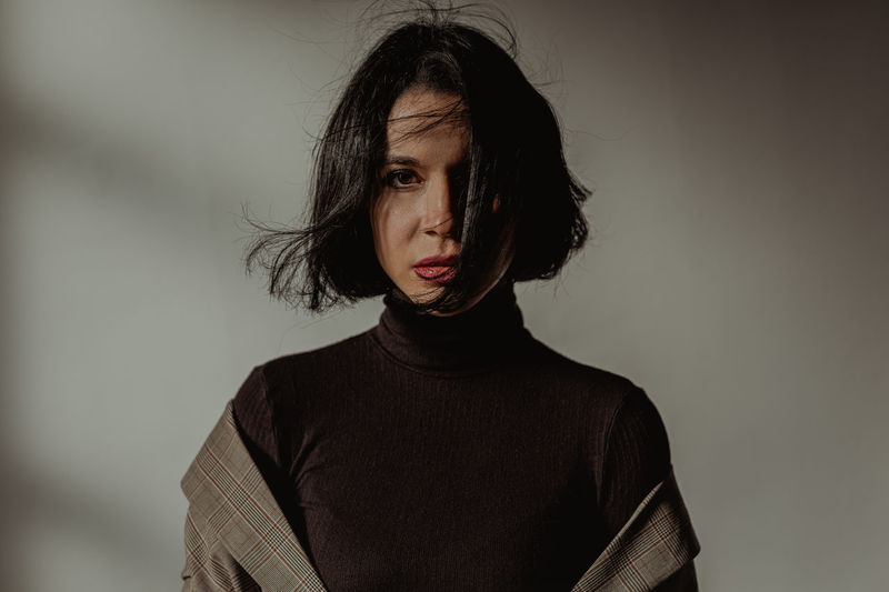 Tranquil female with flying hair and in black turtleneck looking at camera in room against white wall