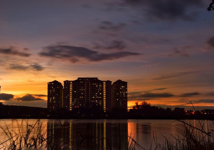 Silhouette buildings by lake against sky during sunset