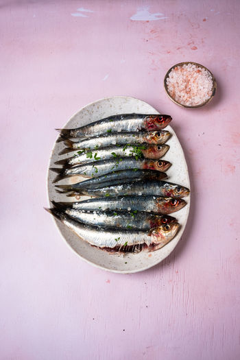 Top view of bunch of fresh sardines placed on oval plate on pink table near bowl with salt