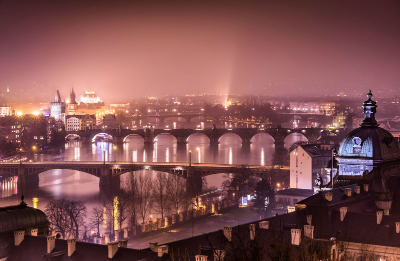 Illuminated bridge over river against buildings in city at night