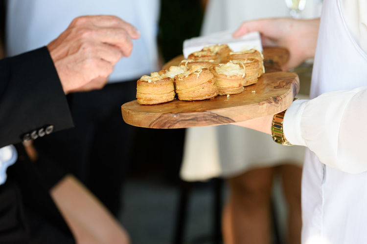 Hand taking appetizer with a cheese filling on a wooden plate hold by a waiter in a white vest