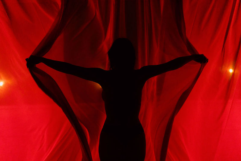 Silhouette of unrecognizable female opening light red curtains with spread arms while standing in murk room at dark night time