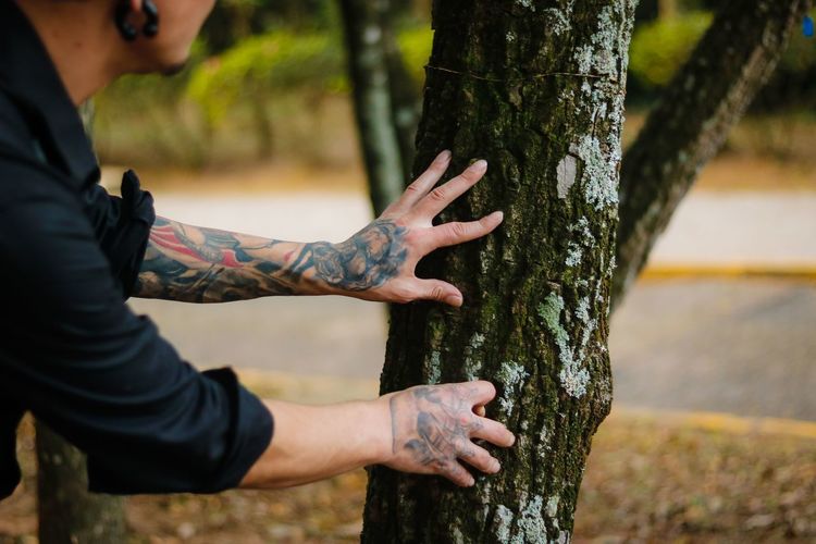 Midsection of man with tattooed hands touching tree trunk at park