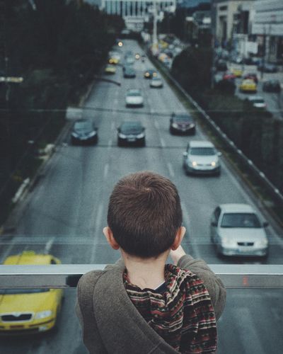 Rear view of boy standing by railing over cars on road in city
