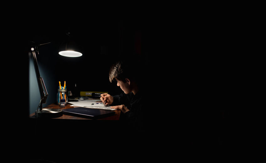 Teenage boy drawing at a desk in a dark room by lamp light.
