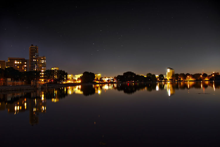 Illuminated buildings in city reflecting on calm lake against sky at night