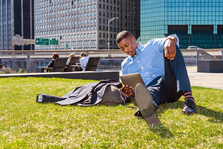 A young black man is sitting on a green lawn in new york city, reading on a tablet computer.