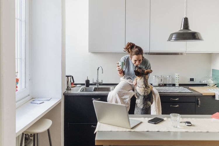 Female professional playing with dog while sitting on kitchen counter during coffee break