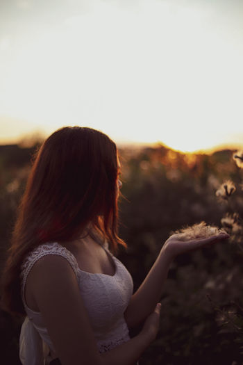 Side view of woman with dandelions standing against sky during sunset