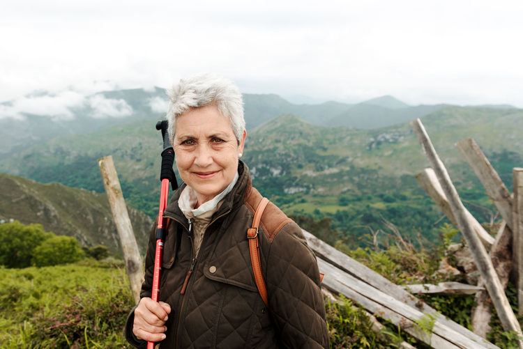 Smiling elderly woman with backpack and trekking stick standing on grassy slope towards mountain peak during trip in nature looking at camera