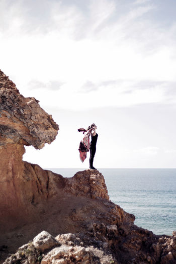 Man jumping on rock by sea against sky