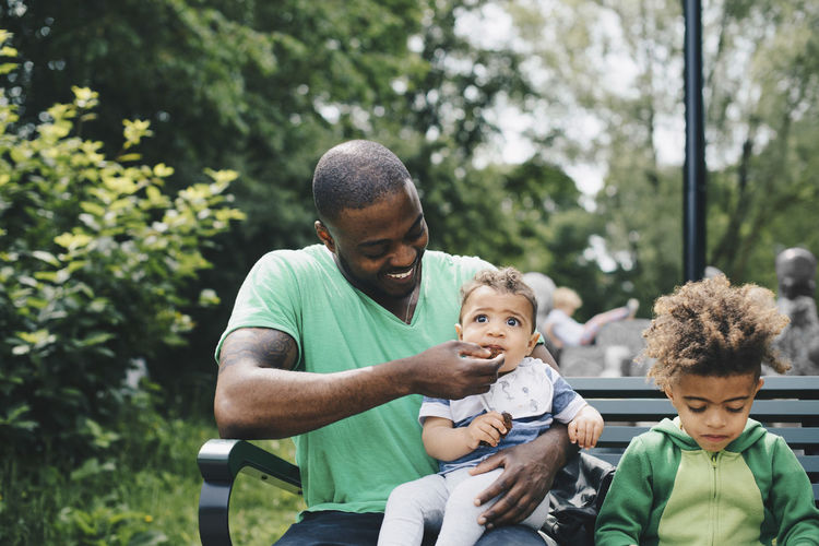 Smiling father feeding baby boy while sitting with son on bench at park