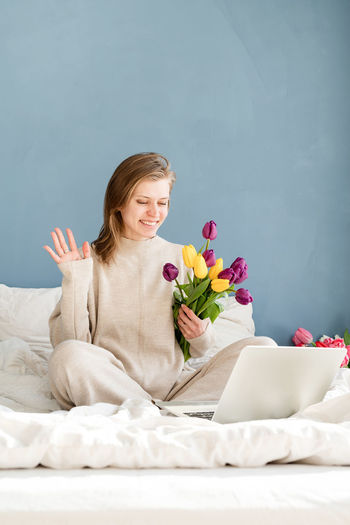 Smiling woman sitting on the bed wearing pajamas, with pleasure enjoying flowers, working on laptop