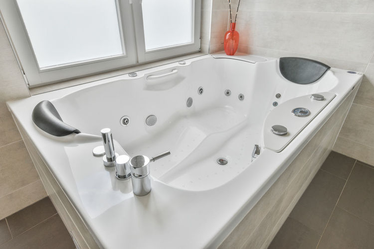 High angle of large white bathtub with hydromassage supplies located near window in modern luxurious bathroom at home