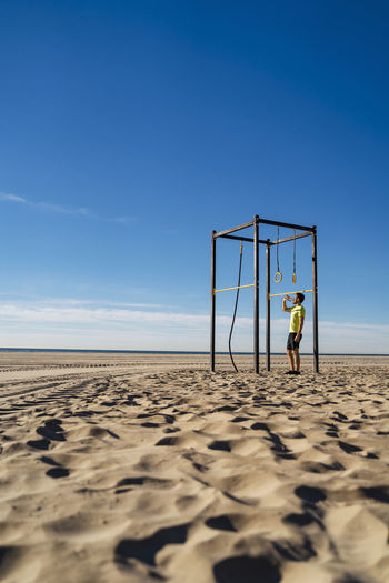 Athlete drinking water standing by gymnastic rings at beach on sunny day