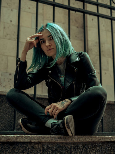 Low angle portrait of young woman with green hair sitting against fence