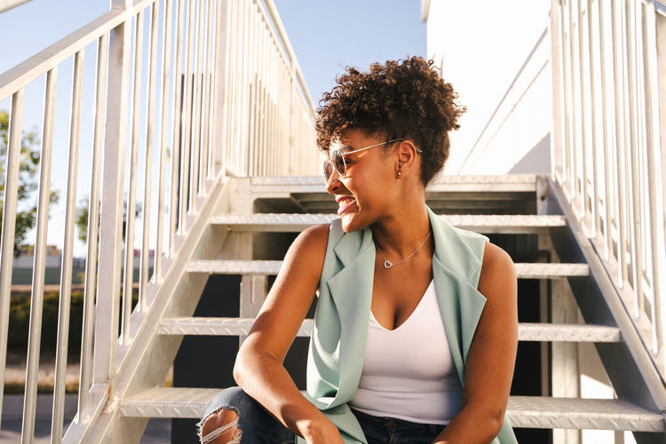 Low angle of cheerful young african american hipster female in trendy outfit and sunglasses sitting on stairs on urban street