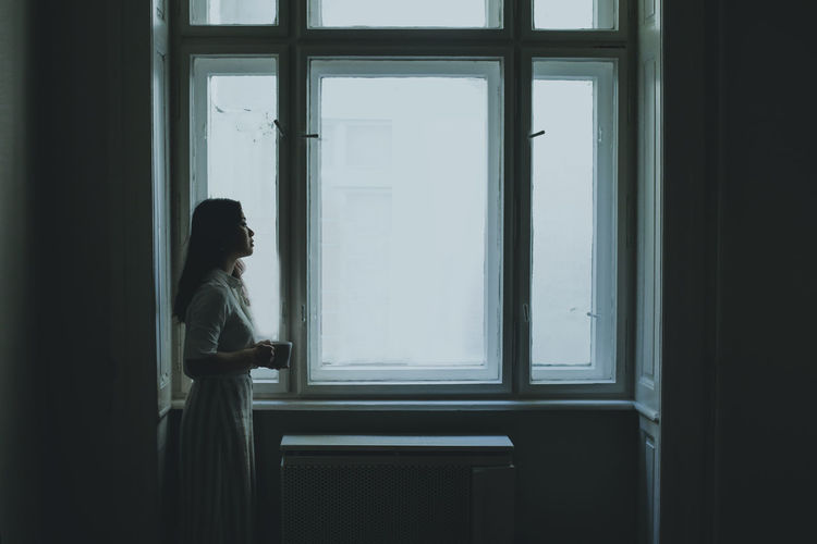 Side view of woman with coffee cup standing by window