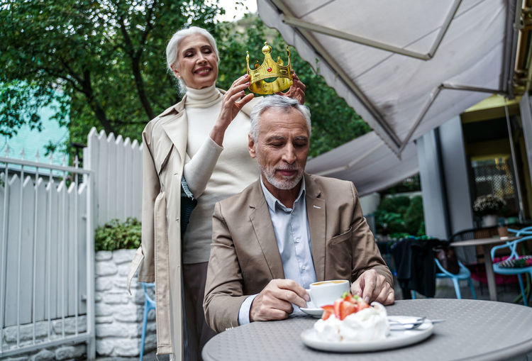 Smiling woman holding crown over man head while sitting at cafe