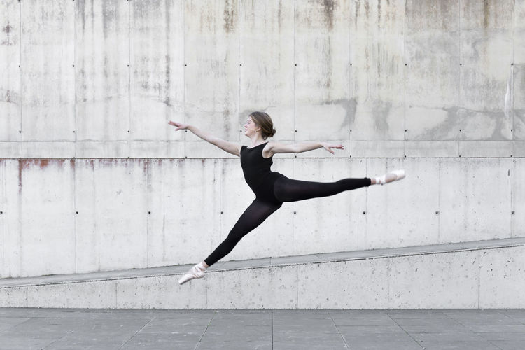 Ballerina in black leotard jumping in front of concrete wall