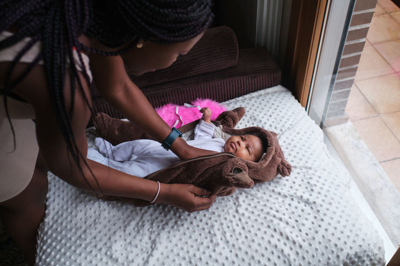 Caring african american mother with long braided hair dressing bodysuit on newborn baby lying on blanket on sofa