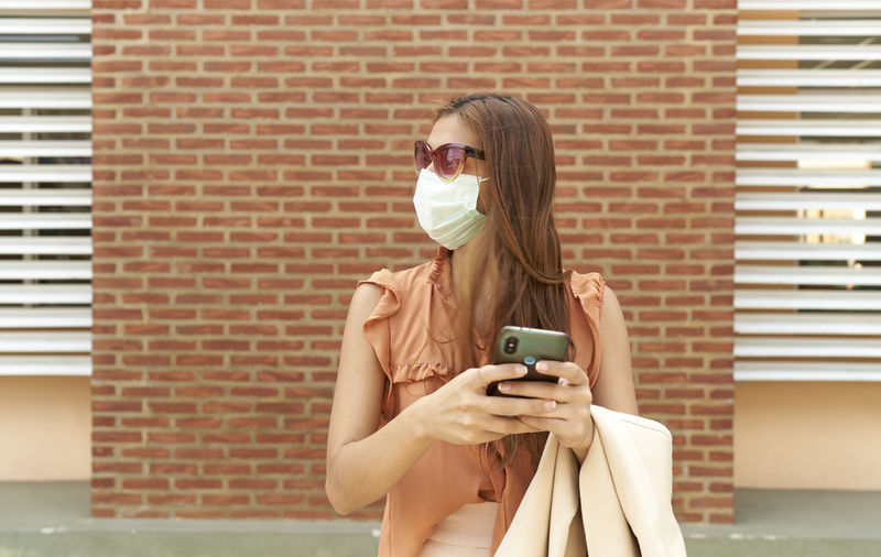 Woman standing on brick wall, young businesswoman with her mask talking with her cell phone