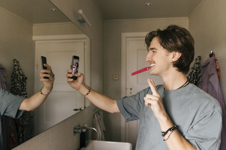Smiling man taking selfie through smart phone while brushing teeth in bathroom at home