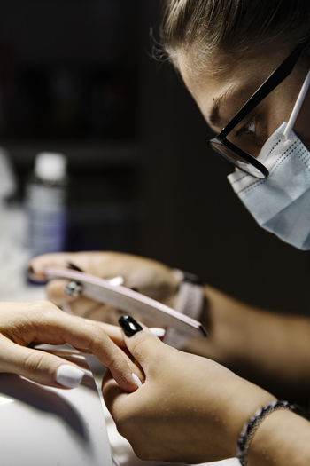 Woman in mask shaping nails of client while working during epidemic