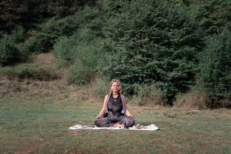 Female sitting in lotus pose on blanket near healing crystals with wireless earphones listening to music and meditating with closed eyes in nature