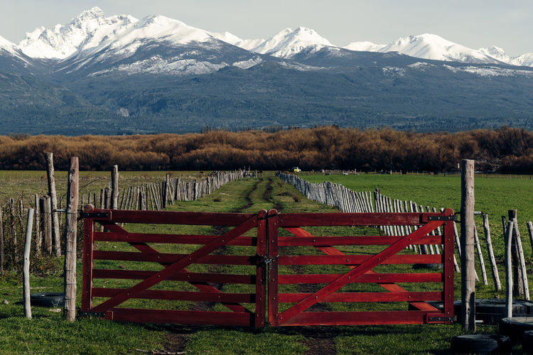 Spectacular scenery of rural road behind shabby wooden fence and gates in valley on background of mountain range covered with snow in patagonia