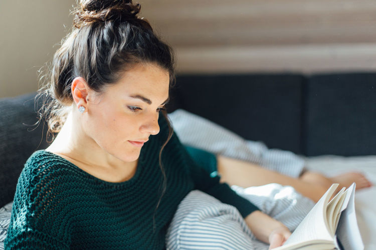 Young woman reading book while lying on bed at home