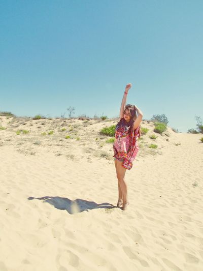Portrait of woman with arm raised tiptoeing on sand against clear blue sky