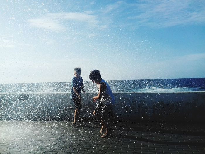 Waves splashing on boys standing on promenade against sky