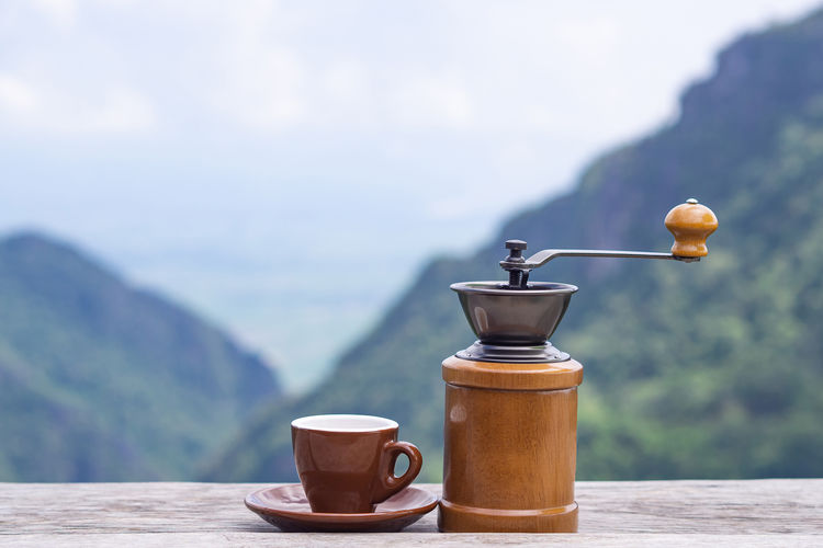 Manual coffee grinder and a coffee cup on wooden table with beautiful scenery view of mountains.