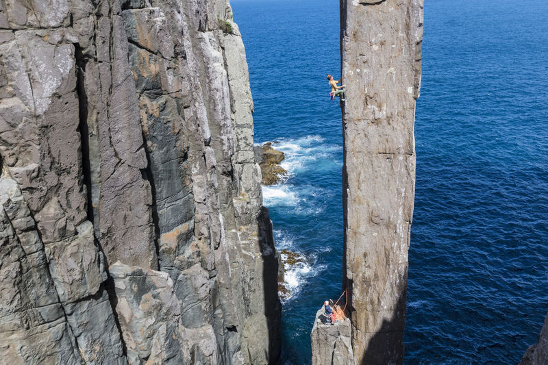 Athletic woman rockclimbs an exposed rock column with her partner belaying her from a ledge, with a sea cliff and the ocean in the backround in the totem pole, cape hauy, tasman national park