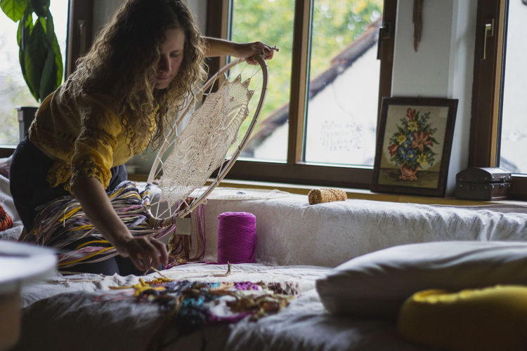 Side view of woman creating handmade dreamcatcher with long threads spending time in house