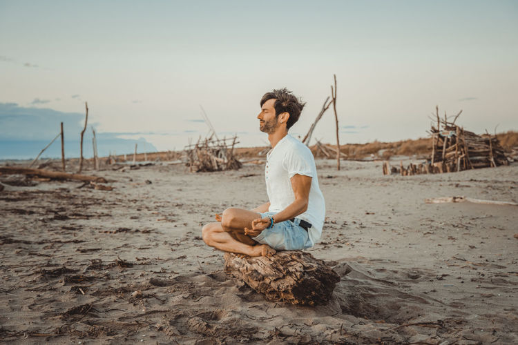 Young man sitting on shore at beach against sky