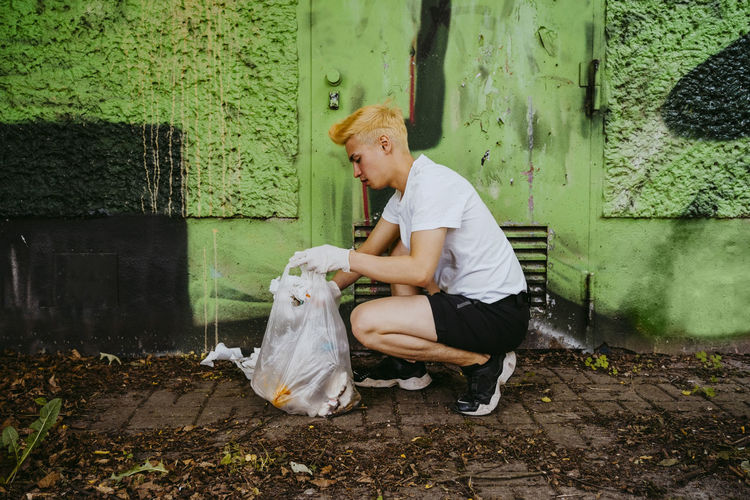 Full length side view of young male volunteer crouching while picking up plastic waste by green wall