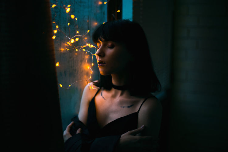 Close-up of woman with illuminated string lights on window at home