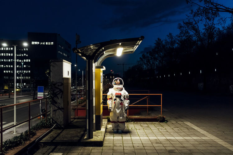 Woman astronaut standing under illuminated light at telephone booth in city during night