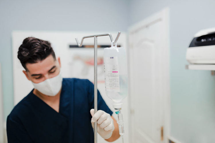 Anonymous young male nurse in respiratory mask installing drop counter with solution in transparent bag in hospital
