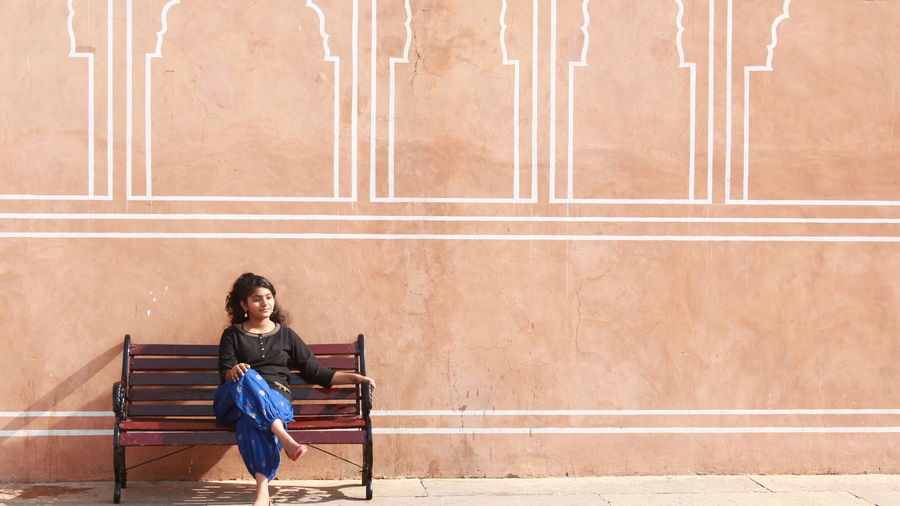 Full length of young woman sitting on bench against wall