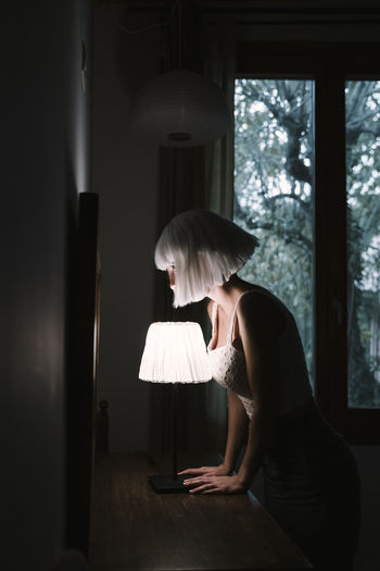 Young woman standing by illuminated lamp at home