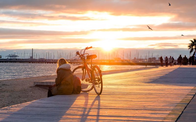 Woman sitting with bicycle at jetty by sea against sky during sunset