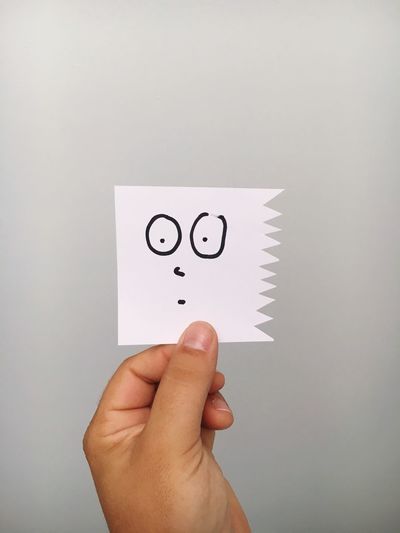 Cropped hand of person holding shocked emotion face on paper against gray background