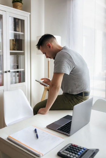 Male entrepreneur using smart phone while sitting on desk by laptop at home office