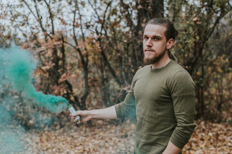 Portrait of young man holding distress flare while standing in forest during autumn