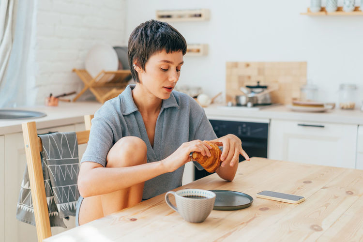 Young european woman sitting in the kitchen eats a croissant and drinks coffee for breakfast.